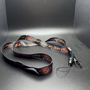 May include: Black lanyard with a repeating pattern of a stylized orange design and the words "EIGHT MAKES ONE TEAM" and "SNODROP". The lanyard has a black plastic clasp and a metal clip.