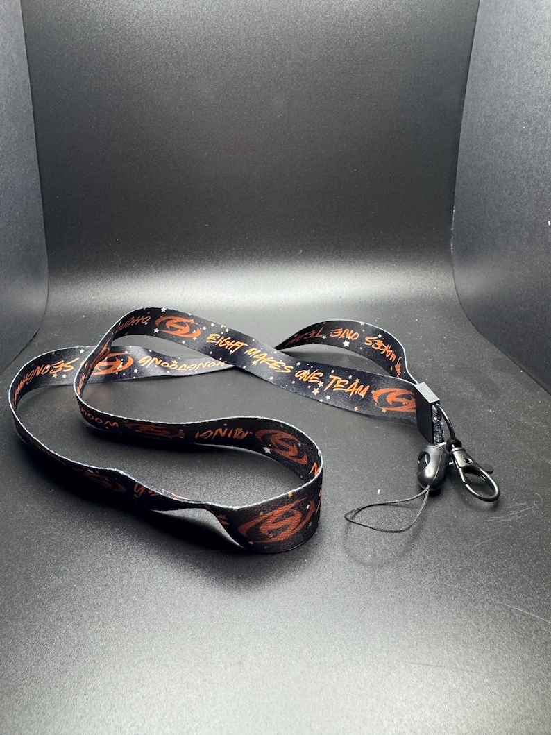 May include: Black lanyard with a repeating pattern of a stylized orange design and the words "EIGHT MAKES ONE TEAM" and "SNODROP". The lanyard has a black plastic clasp and a metal clip.
