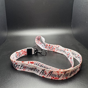 May include: White lanyard with red splatter design and black text that reads "KIDS EVERYWHERE ALL AROUND THE WORLD". The lanyard also features the text "Seungmin Chan" and a black clasp.
