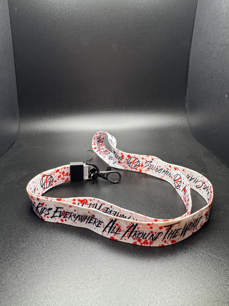 May include: White lanyard with red splatter design and black text that reads "KIDS EVERYWHERE ALL AROUND THE WORLD". The lanyard also features the text "Seungmin Chan" and a black clasp.