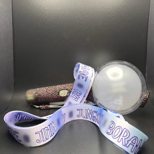 May include: A light stick with a clear, round head and a handle covered in small, sparkling embellishments. A white and blue ribbon with text is draped across the light stick. The background is dark.