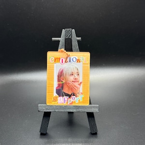 May include: A clear acrylic keychain with a photo of a person with white and pink hair. The keychain has an orange border with the words "emotional support" in colorful text. The keychain is displayed on a small black easel.