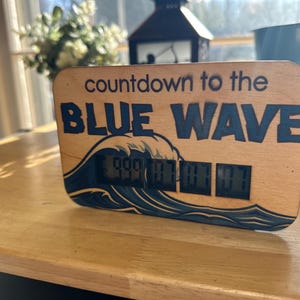 Puede incluir: Temporizador de cuenta regresiva de madera con el texto "countdown to the BLUE WAVE" y un gráfico de olas. La pantalla digital muestra los números 999, 07, 07, 07. El temporizador está sobre una superficie de madera.