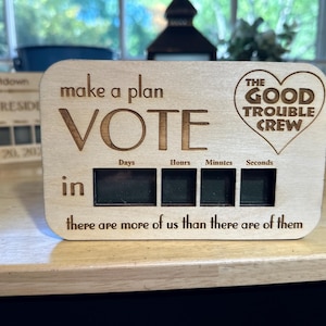 Puede incluir: Un temporizador de cuenta atrás de madera con el texto "make a plan VOTE" y un corazón con el texto "THE GOOD TROUBLE CREW". El temporizador tiene cuatro pantallas digitales para días, horas, minutos y segundos. El texto "in there are more of us than there are of them" está debajo del temporizador.
