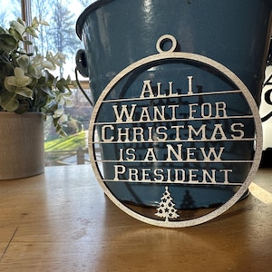 Puede incluir: Adorno navideño redondo plateado con el texto "All I Want For Christmas Is A New President" y un pequeño diseño de árbol de Navidad. El adorno cuelga frente a un cubo azul.