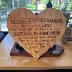Puede incluir: Un letrero de madera en forma de corazón con el texto "En esta casa, creemos: Ningún humano es ilegal, el agua es vida, la ciencia es real, el odio no tiene cabida aquí, los derechos trans son derechos humanos, la diversidad nos hace más fuertes, las vidas negras importan, el amor es amor."
