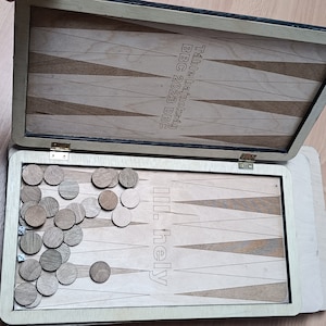 May include: A wooden backgammon board game set with a black and white design. The board is open and the pieces are scattered on the board. The board has the text "The Game That Makes You Think" and "ill. hely" on it.