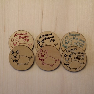 May include: Six round, brown wooden coasters with black pig illustrations and red and blue text. The text on the coasters reads "Emotional Support Pig" and "Dapper Pig thinks you're GREAT!"