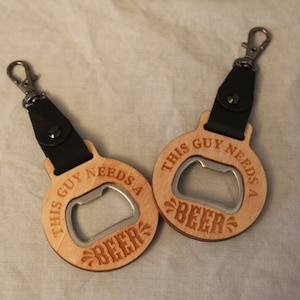 May include: Two wooden bottle openers with black leather straps and metal rings. Each opener has the text "This guy needs a beer" engraved on it.