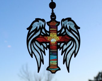 May include: A stained glass window hanging with a colorful cross and angel wings. The cross has a sunset scene with orange, yellow, red, and blue colors. The wings are black and white.