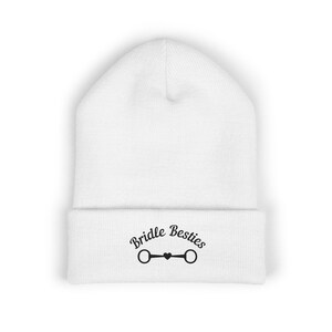 May include: A white knit beanie with a folded cuff. The cuff features black embroidered text that reads Bridle Besties, positioned above a graphic of a horse bit with a small heart in the center, creating a clean and simple design.