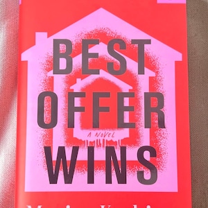 May include: A book titled Best Offer Wins by Marisa Kashino with a bright red and pink cover. The design features a central house graphic with a spray-painted effect and bold dark text. A small circular logo is located in the top right corner.