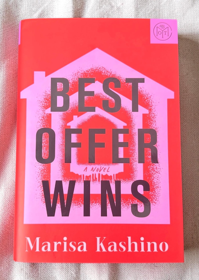 May include: A book titled Best Offer Wins by Marisa Kashino with a bright red and pink cover. The design features a central house graphic with a spray-painted effect and bold dark text. A small circular logo is located in the top right corner.