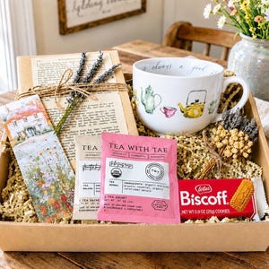 May include: A gift box containing a white ceramic mug with tea pot illustrations, a Tea With Tae herbal tea sachet, a Lotus Biscoff cookie pack, a floral bookmark, and dried lavender stems, all nestled in a cardboard box with paper filler.