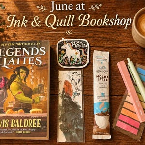 May include: A still-life arrangement featuring a book titled "Legends & Lattes" by Travis Baldree, a coffee cup with latte art, a "Stay Magical" sticker, a bookmark, a mocha latte packet, a pen, and sticky notes. The scene is set on a wooden surface.