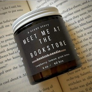 May include: A glass candle jar with a white screw-top lid and a black label. The label features the white text: A SCENT STORY, MEET ME AT THE BOOKSTORE, bookish candle, raspberry, lemon, pink lotus, 4 oz. / 40 hrs. The candle is dark-coloured.