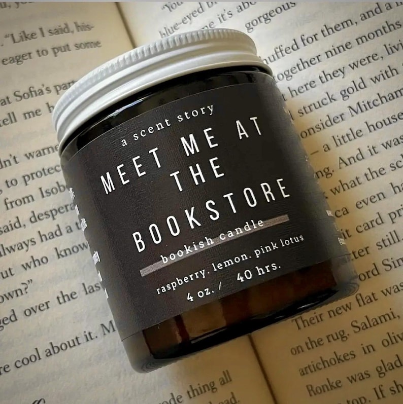 May include: A glass candle jar with a white screw-top lid and a black label. The label features the white text: A SCENT STORY, MEET ME AT THE BOOKSTORE, bookish candle, raspberry, lemon, pink lotus, 4 oz. / 40 hrs. The candle is dark-coloured.