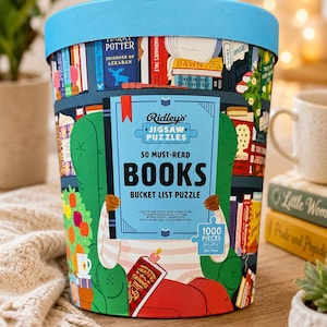 May include: A cylindrical puzzle container with a light blue lid, featuring a colorful illustration of a person reading in a green armchair surrounded by bookshelves. The label reads Ridley's Jigsaw Puzzles 50 Must-Read Books Bucket List Puzzle 1000 Pieces.