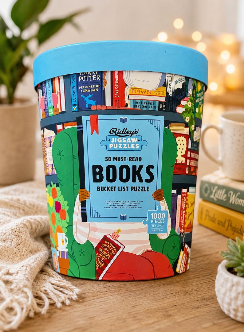 May include: A cylindrical puzzle container with a light blue lid, featuring a colorful illustration of a person reading in a green armchair surrounded by bookshelves. The label reads Ridley's Jigsaw Puzzles 50 Must-Read Books Bucket List Puzzle 1000 Pieces.