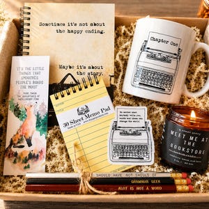 May include: A gift box containing a spiral notebook with the text Sometimes it's not about the happy ending, a white mug with a typewriter illustration, a bookmark with a fox, a yellow memo pad, a typewriter sticker, a candle, and three pencils.