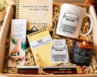 May include: A gift box containing a spiral notebook with the text Sometimes it's not about the happy ending, a white mug with a typewriter illustration, a bookmark with a fox, a yellow memo pad, a typewriter sticker, a candle, and three pencils.