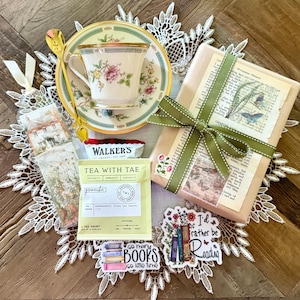 May include: A tea-themed gift set featuring a floral teacup and saucer, a gold spoon, a wrapped book with a green ribbon, tea sachets, and stickers that read "so many books so little time" and "I'd rather be reading."