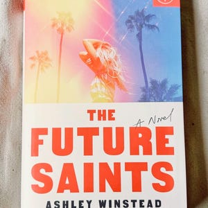 May include: A paperback book titled The Future Saints by Ashley Winstead. The cover features a person with long hair standing against a sunset sky with palm trees, rendered in gradients of yellow, pink, and blue. The title is in bold red text.