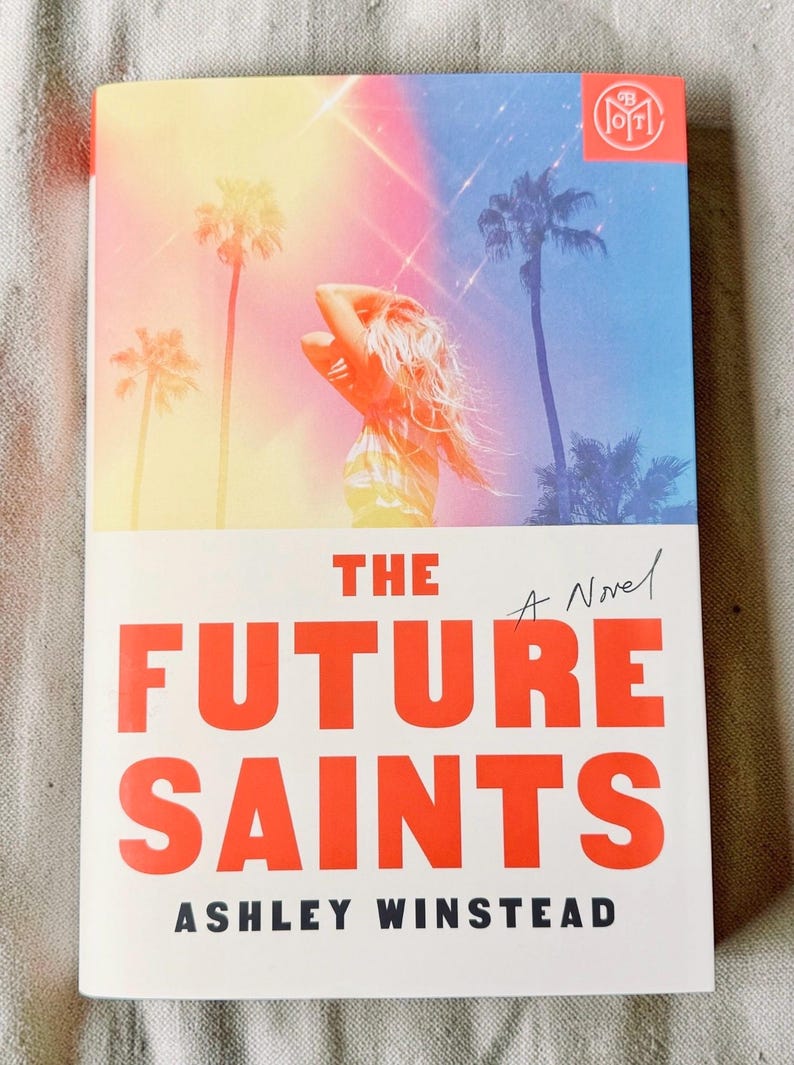 May include: A paperback book titled The Future Saints by Ashley Winstead. The cover features a person with long hair standing against a sunset sky with palm trees, rendered in gradients of yellow, pink, and blue. The title is in bold red text.