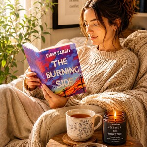 May include: A paperback book titled The Burning Side by Sarah Damoff with a colourful sunset cover, a ceramic mug with blue floral patterns, a lit amber glass candle labelled Meet Me At The Bookstore, and a small plate with chocolate squares.
