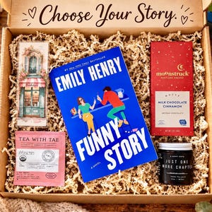 May include: A gift box with the text Choose Your Story containing a blue book titled Funny Story by Emily Henry, a Moonstruck milk chocolate cinnamon bar, a Tea With Tae tea bag, a bookstore bookmark, and a Just One More Chapter candle.