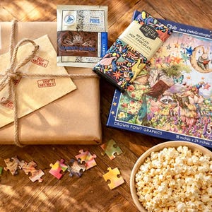 May include: A gift set featuring a brown paper-wrapped package tied with twine, a Harney & Sons Paris tea sachet, a Seattle Chocolate Mexican Hot Chocolate bar, a 500-piece Crown Point Graphics puzzle box, and a bowl of popcorn with loose puzzle pieces.