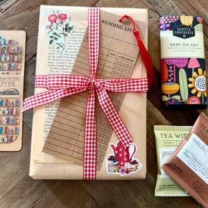 May include: A gift set featuring a book wrapped in brown paper with a red and white gingham ribbon, a reading list card, a bookmark with a bookshelf print and blue tassel, a Seattle Chocolate dark sea salt bar, and two Tea With Tae tea sachets.