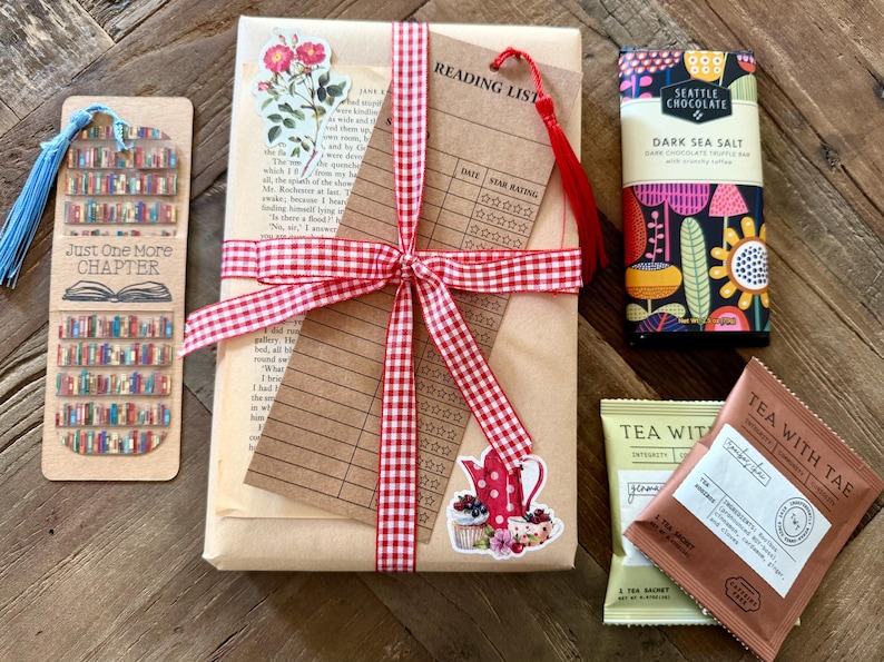 May include: A gift set featuring a book wrapped in brown paper with a red and white gingham ribbon, a reading list card, a bookmark with a bookshelf print and blue tassel, a Seattle Chocolate dark sea salt bar, and two Tea With Tae tea sachets.