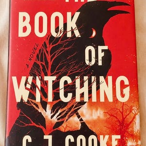May include: A hardback book titled The Book of Witching by C. J. Cooke. The cover features a large black raven silhouette against a vibrant red background, with a bare red tree and the text: Four hundred years separate them. One book binds them.