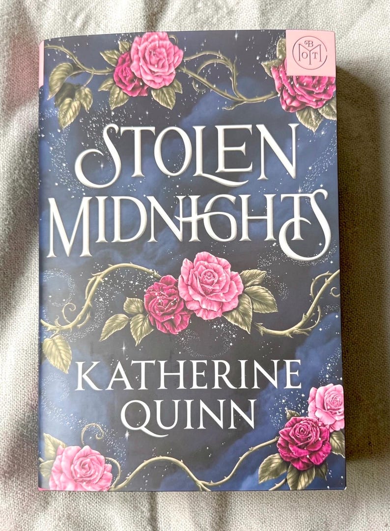 May include: A book titled Stolen Midnights by Katherine Quinn with a dark blue, starry night sky background. The cover features pink roses on thorny vines, a small pink square logo in the top right corner, and white serif lettering.