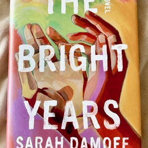 May include: A book titled The Bright Years by Sarah Damoff with a cover illustration featuring two hands reaching upward against a colourful background of orange, yellow, pink, and green. The title is printed in large white letters across the centre.