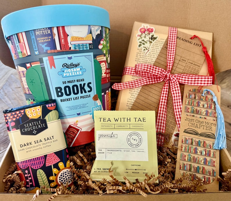 May include: A gift box containing a Ridley's 50 Must-Read Books jigsaw puzzle, a Seattle Chocolate Dark Sea Salt truffle bar, a Tea With Tae genmaicha sachet, a book-themed reading list card with a red gingham ribbon, and a Just One More Chapter bookmark.