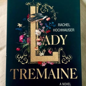 May include: A hardback book titled Lady Tremaine by Rachel Hochhauser. The dark green cover features a large, gold-textured letter L intertwined with colourful floral illustrations and a bird. The title and author name are printed in gold lettering.