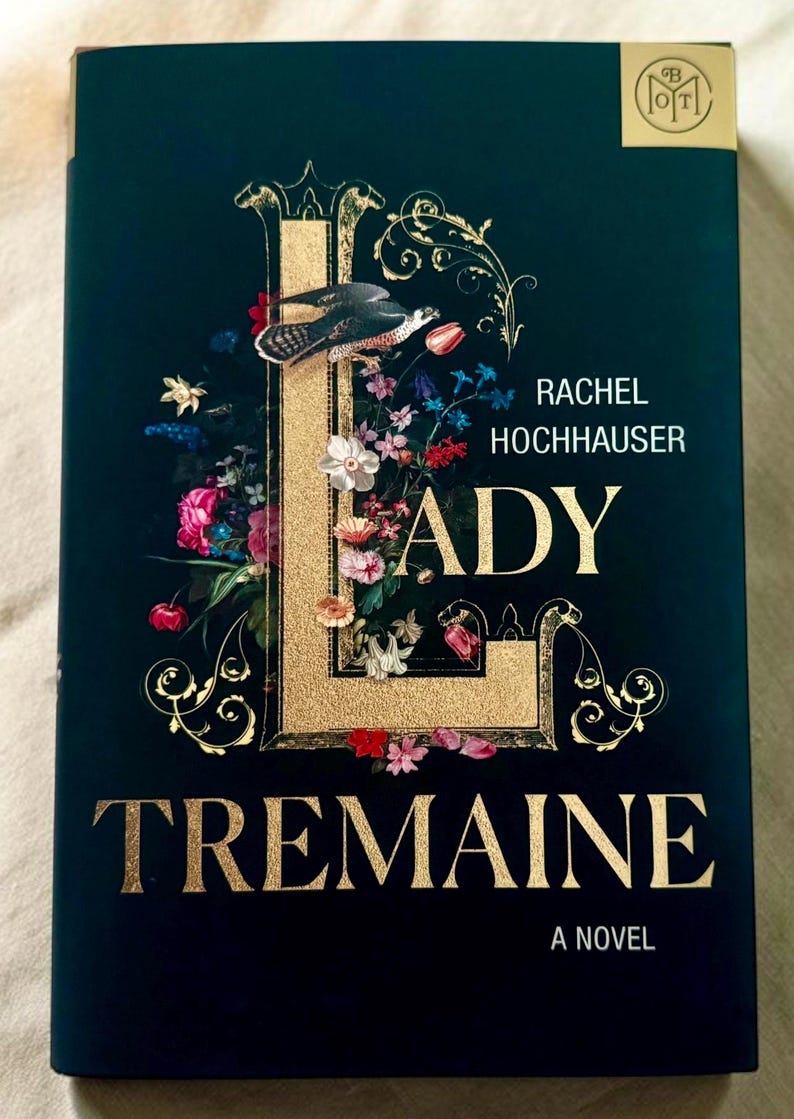 May include: A hardback book titled Lady Tremaine by Rachel Hochhauser. The dark green cover features a large, gold-textured letter L intertwined with colourful floral illustrations and a bird. The title and author name are printed in gold lettering.