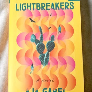 May include: A book titled Lightbreakers by Aja Gabel with a yellow cover featuring a green cactus, two small figures, and two birds. The background has a repeating pattern of overlapping pink and orange circles, with a teal logo in the top corner.