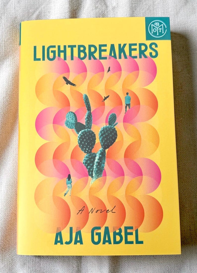 May include: A book titled Lightbreakers by Aja Gabel with a yellow cover featuring a green cactus, two small figures, and two birds. The background has a repeating pattern of overlapping pink and orange circles, with a teal logo in the top corner.