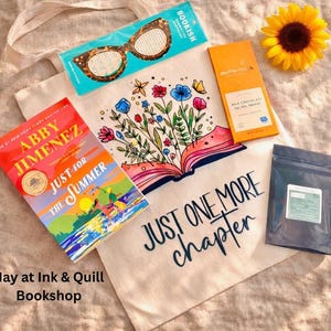 May include: A flat lay featuring a canvas tote bag with the text "JUST ONE MORE chapter" and a floral book design. The image includes a book, chocolate bar, sunflower, and a bag of tea. The book is titled "JUST FOR THE SUMMER".