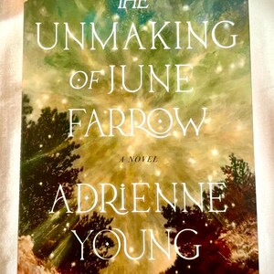 May include: A hardback book titled The Unmaking of June Farrow by Adrienne Young. The cover features a green and gold abstract background with glowing light orbs, dark tree silhouettes at the bottom, and a small gold logo in the top corner.
