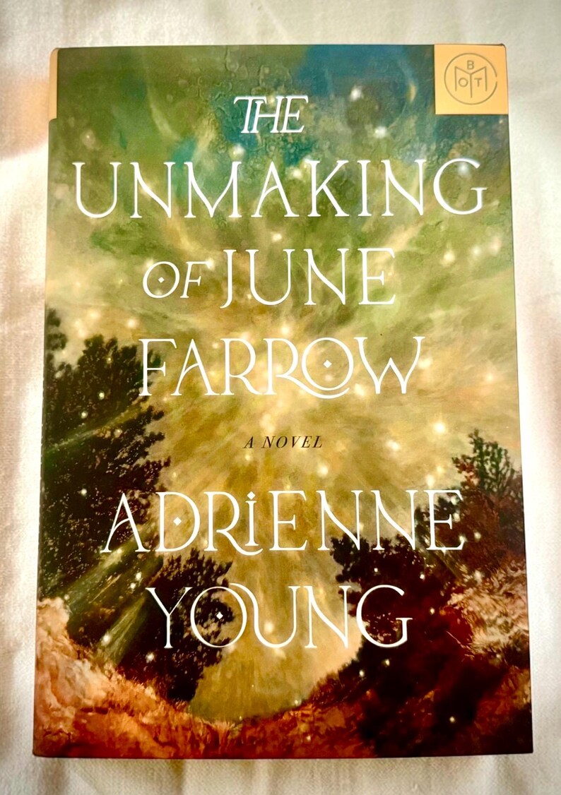 May include: A hardback book titled The Unmaking of June Farrow by Adrienne Young. The cover features a green and gold abstract background with glowing light orbs, dark tree silhouettes at the bottom, and a small gold logo in the top corner.