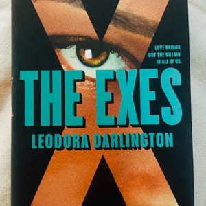 May include: A book titled The Exes by Leodora Darlington with a black cover featuring a large orange letter X, a close-up of a human eye, and the text Love brings out the villain in all of us. The title is in large teal letters on the front.