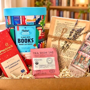 May include: A gift box containing a Ridley's 50 Must-Read Books jigsaw puzzle, a Moonstruck milk chocolate cinnamon bar, a Tea With Tae herbal tea sachet, a book-themed decorative item tied with twine and dried lavender, and a bookmark.