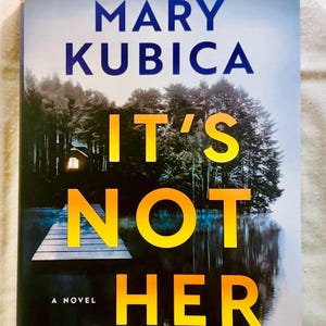May include: A book titled It's Not Her by Mary Kubica. The cover features a dark forest and a wooden dock extending into a lake at night, with a single glowing window visible in the trees. The title text is in large yellow and orange letters.