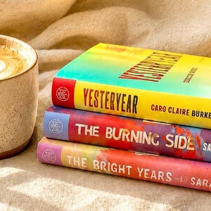 May include: A ceramic mug filled with coffee featuring latte art, positioned next to a stack of three books. The top book is titled Yesteryear by Caro Claire Burke, the middle is The Burning Side by Sarah Damoff, and the bottom is The Bright Years.