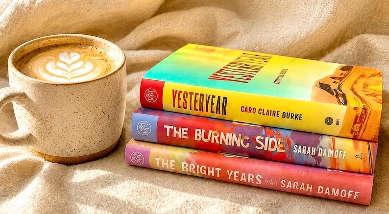 May include: A ceramic mug filled with coffee featuring latte art, positioned next to a stack of three books. The top book is titled Yesteryear by Caro Claire Burke, the middle is The Burning Side by Sarah Damoff, and the bottom is The Bright Years.