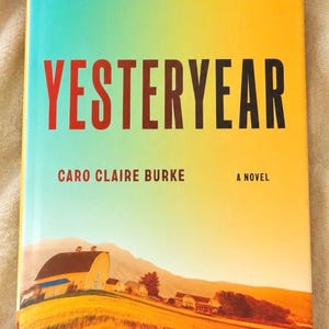 May include: A book titled Yesteryear by Caro Claire Burke with a cover featuring a gradient from teal to yellow. The cover displays a rural landscape with a yellow barn, fields, and mountains, and a red logo in the top right corner.